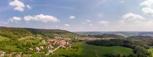Panoramaluftbildaufnahme von Ilmnitz: Im Vordergrund ist die Siedlung mit Häusern umgeben von grünen Ackerflächen, Waldflächen und Bergen