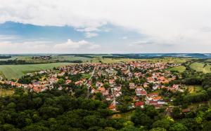 Panoramaluftbild von Cospeda: im Vordergrund grüne Waldflächen gefolgt von der Siedlung mit Häusern, dann folgen grüne Ackerflächen und kleine Waldflächen