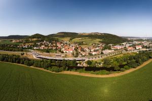 Panoramaluftbild von Göschwitz, im Vordergrund grüne Ackerflächen gefolgt von dem Fluss Saale, dann folgt die Siedlung mit Häusern und im Hintergrund sieht man den alten Steinbruch.