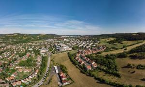 Luftaufnahme von Winzerla: im Vordergrund ist auf der linken Seite der Ortsteil Jena-Süd mit Einfamilienhäusern zu sehen, auf der rechten Seite sind Wiesen, der Hahnengrund mit neuer Wohnsiedlung, gefolgt von Mehrfamilienhäusern, mittig vom Bild ist das ehemalige Heizkraftwerk mit großem Schornstein, gefolgt von dem Ortsteil Neulobeda im Hintergrund mittig, auf der rechten Seite befinden sich die Kernberge und auf der linken Seite sind grüne Waldflächen