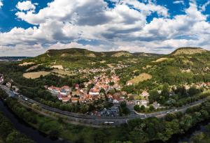 Luftbildaufnahme von Wöllnitz entlang der Saale: Im Vordergrund ist der Fluss, gefolgt von der Schnellstraße Stadtrodaer Straße, gefolgt von der Siedlung mit Häusern, welche sich weit nach hinten ins Gebirge zieht, die Siedlung ist ist umgeben von grünen landwirtschaftlichen Flächen und Wiesen, auf der linken Seite ist der Johannisberg, mittig vom Bild im Hintergrund der Fürstenbrunnen, und links im Bild ist der Ziegenberg