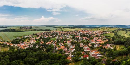 Panoramaluftbild von Cospeda: im Vordergrund grüne Waldflächen gefolgt von der Siedlung mit Häusern, dann folgen grüne Ackerflächen und kleine Waldflächen