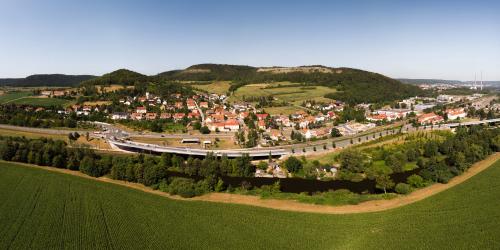 Panoramaluftbild von Göschwitz, im Vordergrund grüne Ackerflächen gefolgt von dem Fluss Saale, dann folgt die Siedlung mit Häusern und im Hintergrund sieht man den alten Steinbruch.