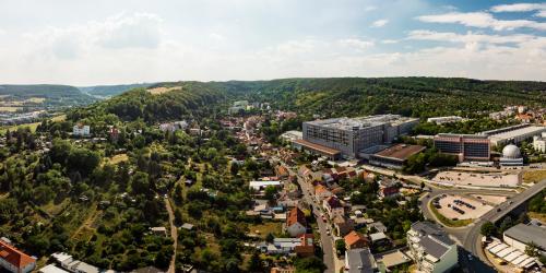 Luftbildaufnahme von Lichtenhain: in der Mitte des Bildes sieht man die Siedlung mit Häusern, welche sich wie eine Art Allee angeordnet sind und am Ende von der Ammerbacher Platte umgeben ist. Rechts im Bild sieht man das Firmengelände von Zeiss. Links im Bild Kleingärten mit viel grün.