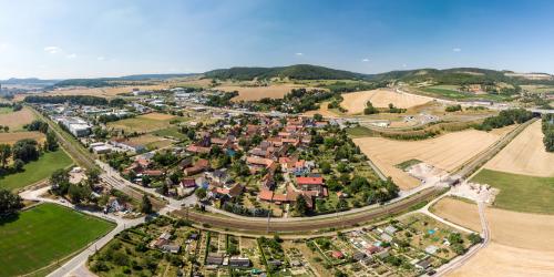 Luftbildaufnahme Maua: Im Vordergrund sind landwirtschaftliche Nutzflächen, gefolgt von einer Kleingartenanlage, dahinter Bahngleisen, gefolgt von der Siedlung mit Häusern, auf der linken Seite ist das Gewerbegebiet und auf der rechten Seite sind landwirtschaftliche Nutzflächen sowie die Autobahnauffahrt A4 Richtung Dresden oder Frankfurt/Main, im Hintergrund ist der Ortsteil Leutra sowie eine bergige Landschaft