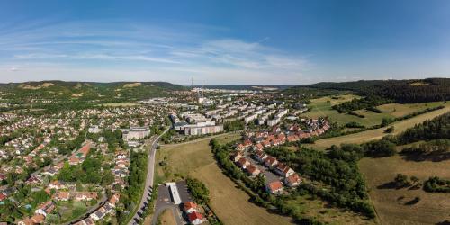 Luftaufnahme von Winzerla: im Vordergrund ist auf der linken Seite der Ortsteil Jena-Süd mit Einfamilienhäusern zu sehen, auf der rechten Seite sind Wiesen, der Hahnengrund mit neuer Wohnsiedlung, gefolgt von Mehrfamilienhäusern, mittig vom Bild ist das ehemalige Heizkraftwerk mit großem Schornstein, gefolgt von dem Ortsteil Neulobeda im Hintergrund mittig, auf der rechten Seite befinden sich die Kernberge und auf der linken Seite sind grüne Waldflächen