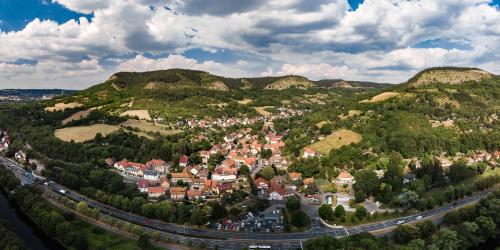 Luftbildaufnahme von Wöllnitz entlang der Saale: Im Vordergrund ist der Fluss, gefolgt von der Schnellstraße Stadtrodaer Straße, gefolgt von der Siedlung mit Häusern, welche sich weit nach hinten ins Gebirge zieht, die Siedlung ist ist umgeben von grünen landwirtschaftlichen Flächen und Wiesen, auf der linken Seite ist der Johannisberg, mittig vom Bild im Hintergrund der Fürstenbrunnen, und links im Bild ist der Ziegenberg