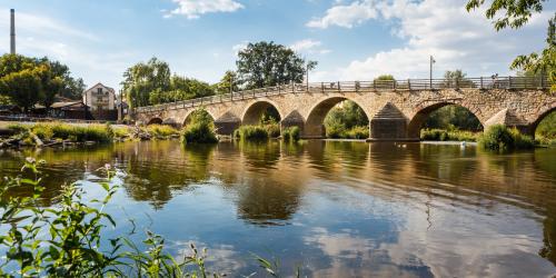 Aufnahme von der Burgauer Brücke am Wehr: Im Vordergrund ist der Fluss die Saale, gefolgt von der historischen Natursteinbrücke mit Gewölbe